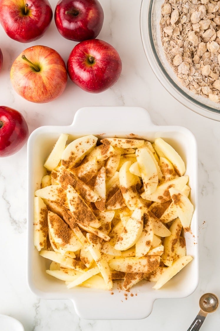 Sliced apples sprinkled with cinnamon in a white baking dish, surrounded by whole apples and a bowl of oat crumb topping on a marble countertop.