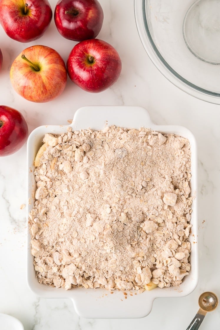 A white baking dish filled with an unbaked apple crumble sits on a marble countertop, surrounded by several whole red apples and a clear mixing bowl.