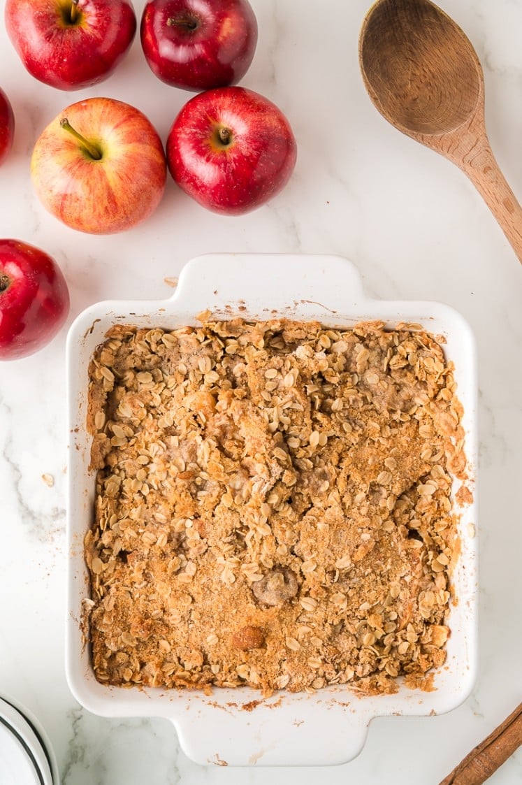 A square white baking dish filled with apple crisp topped with oat crumble sits on a marble countertop, surrounded by whole red apples and a wooden spoon.