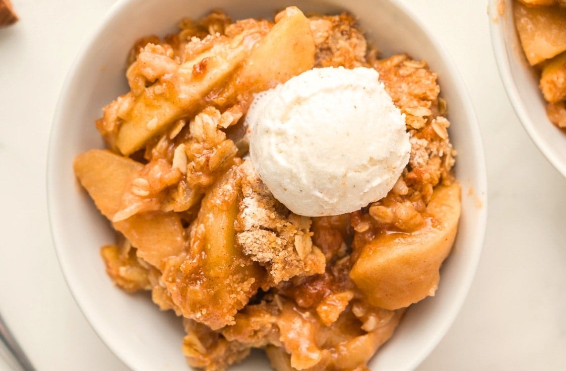 A bowl of apple crisp topped with a scoop of vanilla ice cream, featuring baked apple slices and crumbly oat topping.