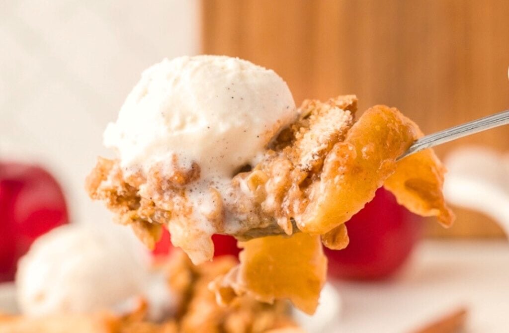 A close-up of a spoonful of apple crumble topped with a scoop of vanilla ice cream, with sliced apples and crumble visible, and a blurred background featuring red apples.