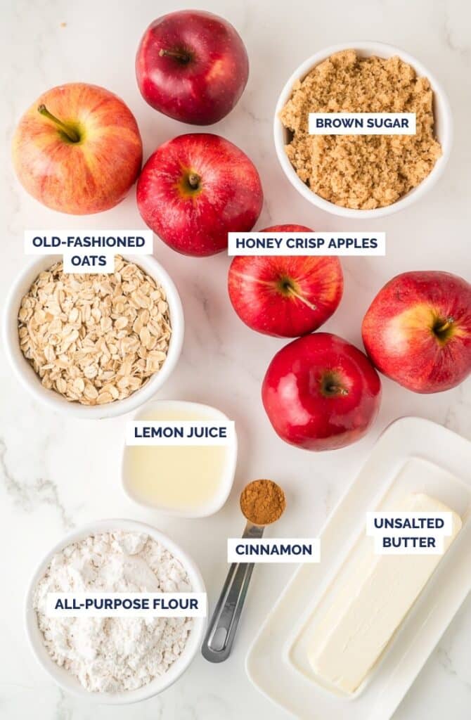 Top-down view of labeled ingredients for apple crisp, including Honeycrisp apples, brown sugar, old-fashioned oats, lemon juice, cinnamon, all-purpose flour, and unsalted butter, arranged on a white surface.