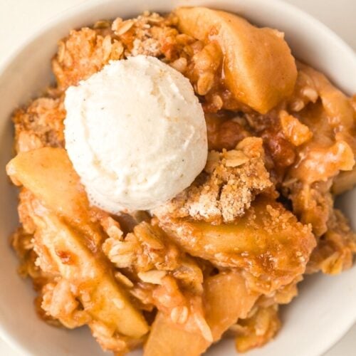 A bowl of apple crisp topped with a scoop of vanilla ice cream, featuring baked apple slices and crumbly oat topping.