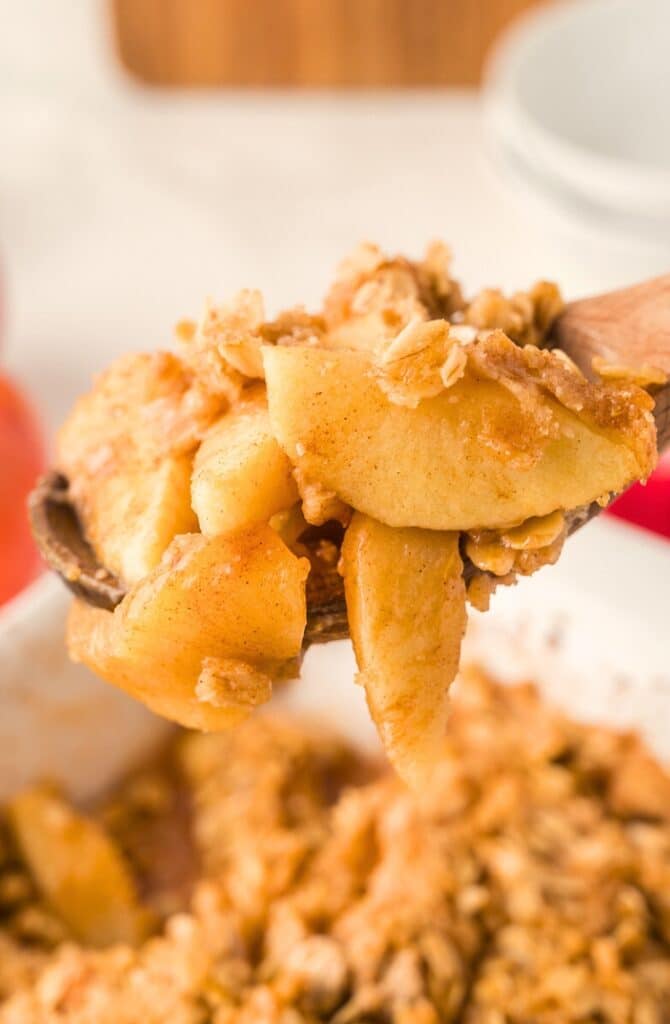 A close-up of a spoonful of apple crisp, showing tender baked apple slices coated in cinnamon and topped with a crumbly oat mixture, with more apple crisp in a dish in the background.