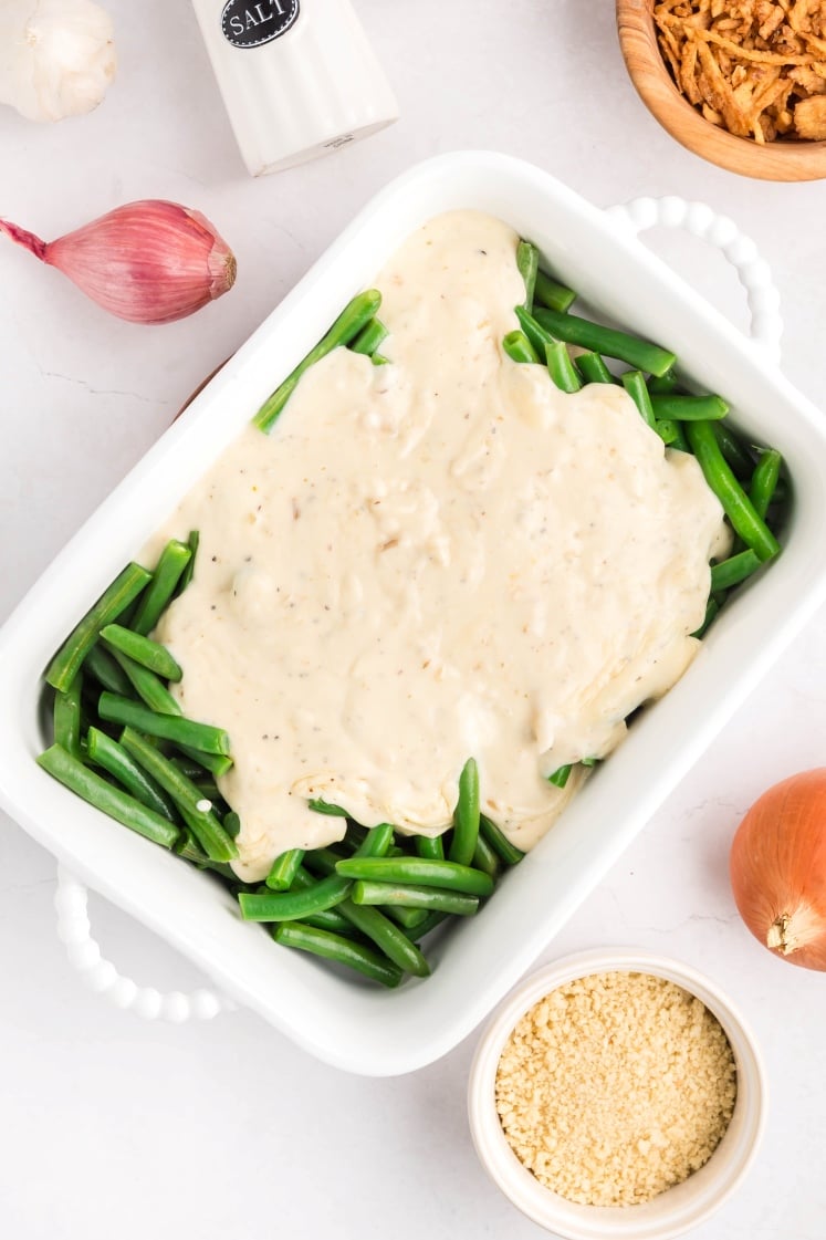 A white baking dish filled with fresh green beans topped with creamy sauce. Surrounding the dish are a shallot, a garlic bulb, a salt shaker, crispy fried onions, an onion, and a bowl of breadcrumbs.