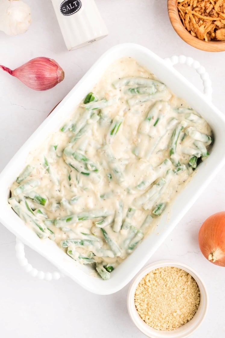 A white baking dish filled with green bean casserole in a creamy sauce, surrounded by a bowl of breadcrumbs, an onion, a shallot, garlic, a salt shaker, and a bowl of crispy fried onions on a white surface.