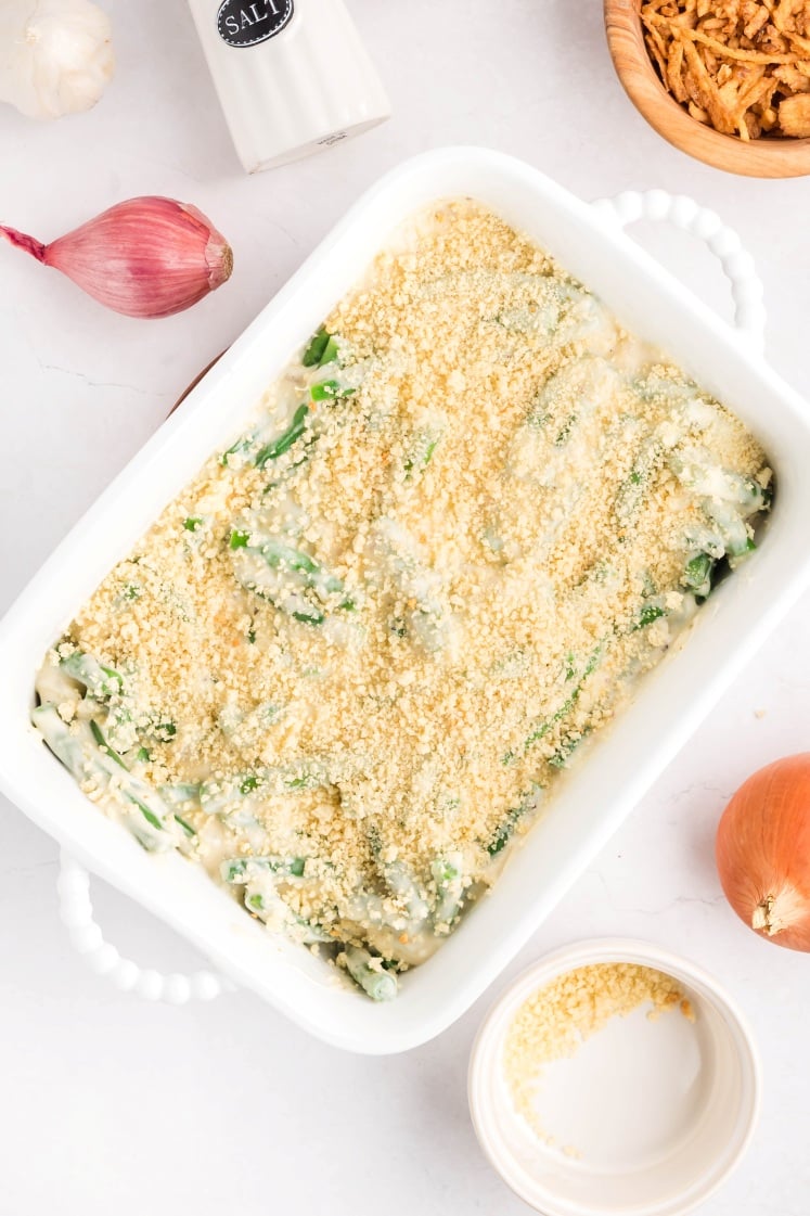 A white baking dish filled with an unbaked green bean casserole topped with breadcrumbs sits on a white countertop, surrounded by a bowl of fried onions, an onion, a shallot, garlic, and a salt shaker.