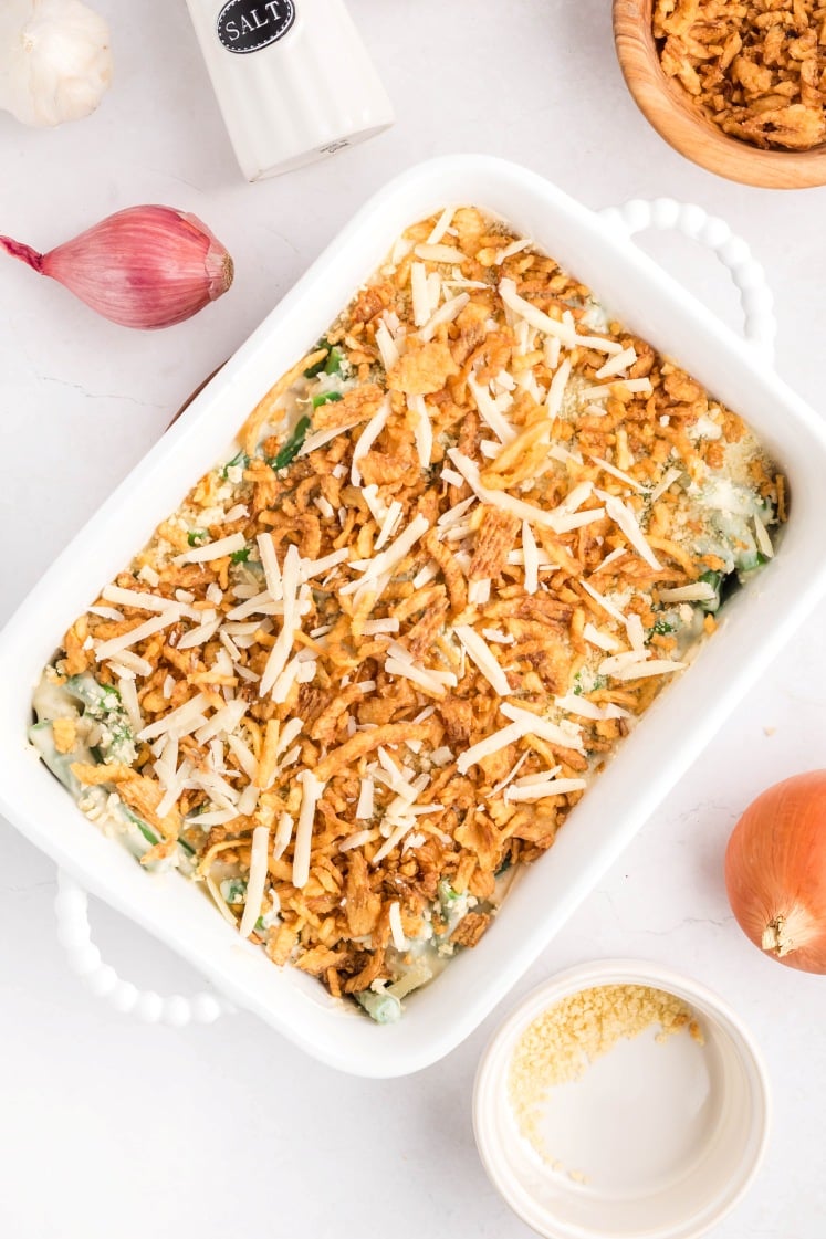 A white baking dish filled with a casserole topped with crispy fried onions and shredded cheese, surrounded by a shallot, onion, garlic, salt shaker, a bowl of fried onions, and a small dish of grated cheese.