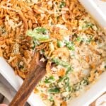 A wooden spoon lifts a serving of creamy green bean casserole topped with crispy fried onions from a white baking dish. The casserole appears baked and golden, with a portion already served.