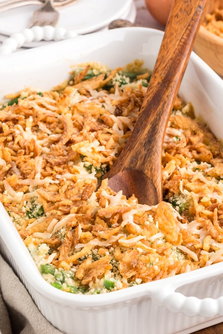 A white casserole dish filled with green bean casserole, topped with crispy fried onions and breadcrumbs, with a wooden serving spoon resting inside the dish.