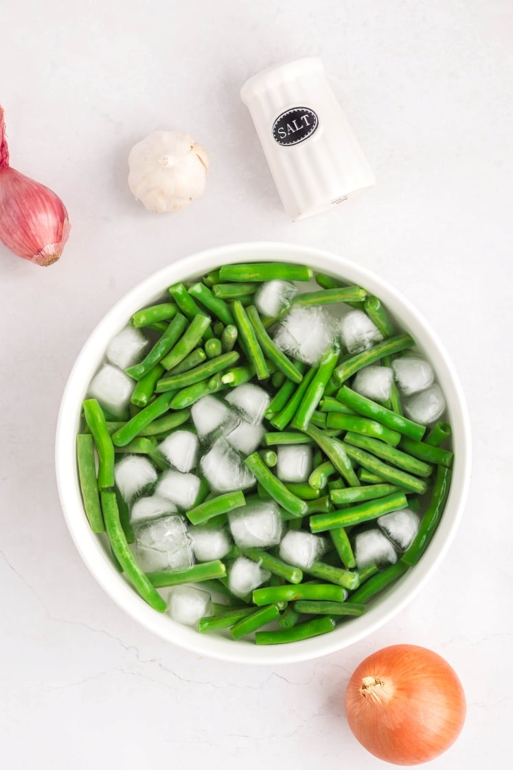 A white bowl filled with green beans and ice cubes sits on a white surface. Nearby are a salt shaker, a garlic bulb, a shallot, and an onion.