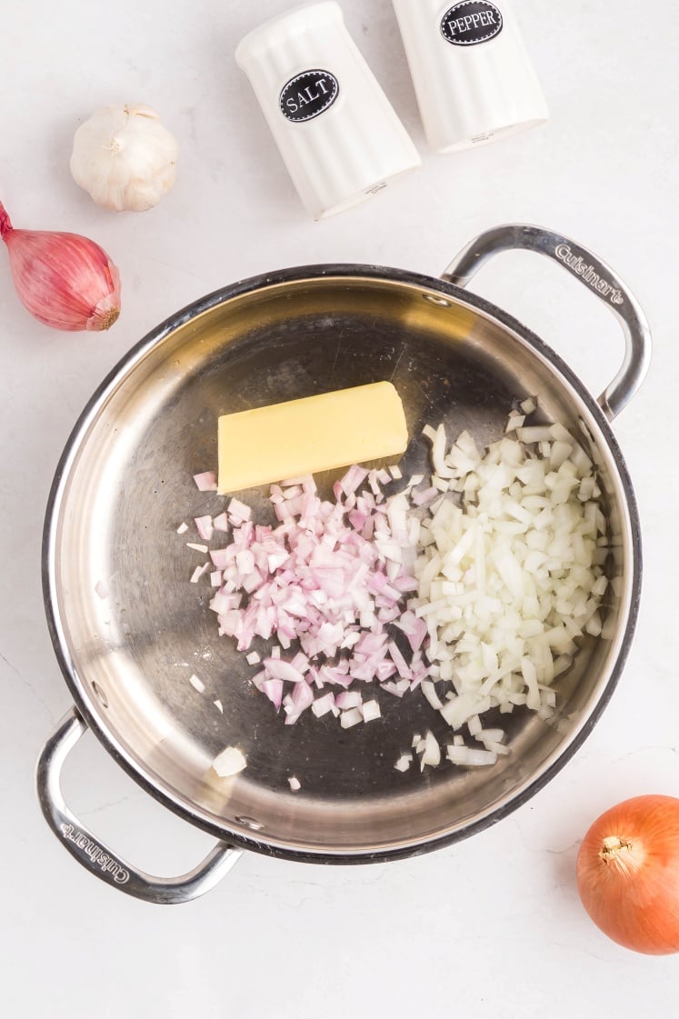 A metal pot contains a stick of butter, chopped onions, and chopped shallots. Nearby on the white surface are a whole shallot, a head of garlic, an onion, and salt and pepper shakers.