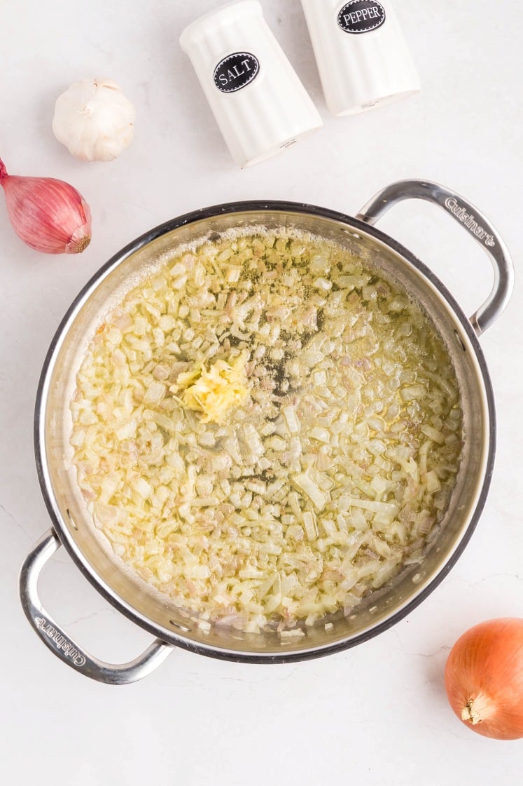 A stainless steel pot filled with sautéed chopped onions and garlic on a white countertop, with a shallot, a whole garlic bulb, an onion, and salt and pepper shakers nearby.