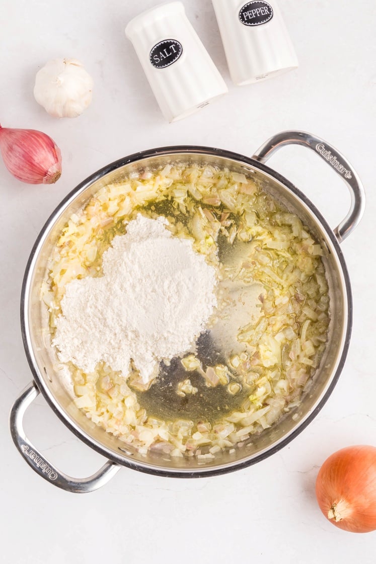A stainless steel pan with sautéed onions and garlic, topped with a mound of flour. Surrounding the pan are a shallot, an onion, a garlic bulb, and salt and pepper shakers on a white surface.
