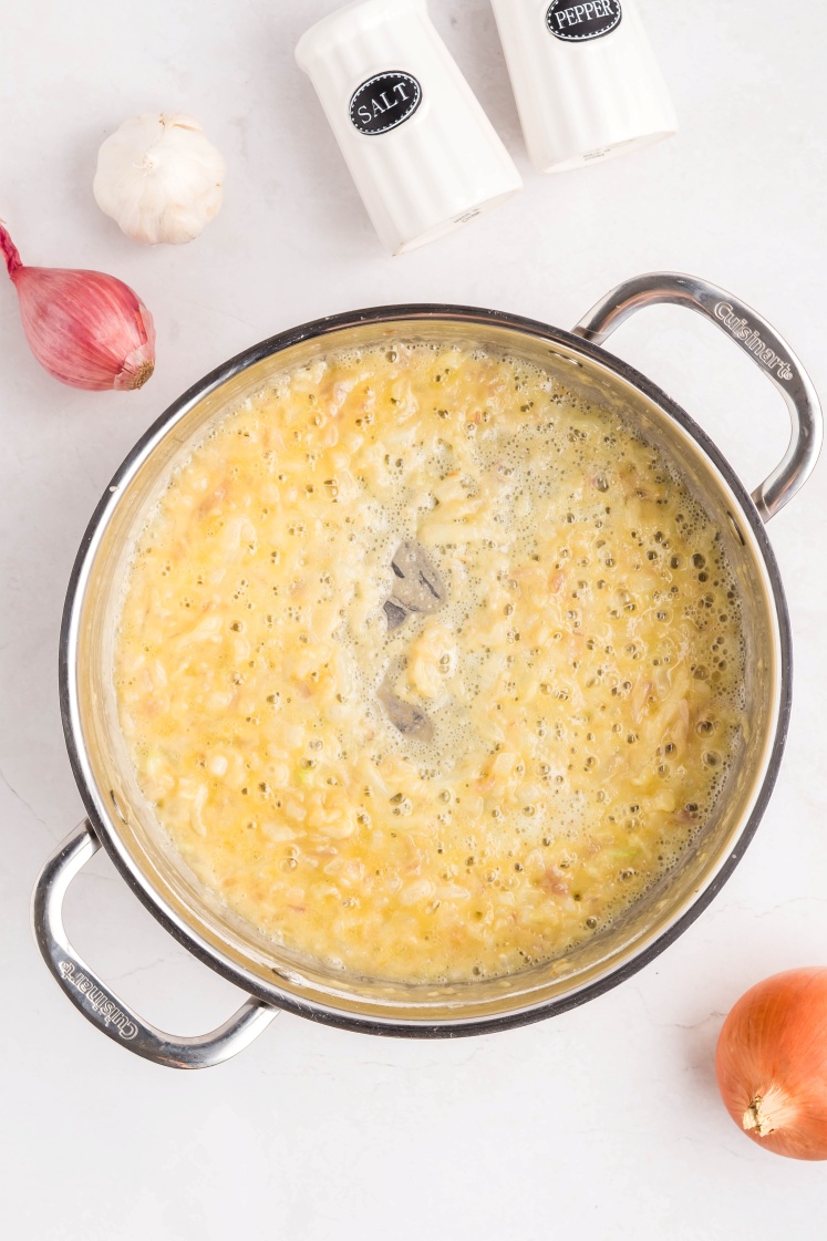 A stainless steel pot with a bubbling yellow mixture, likely a sauce or soup, sits on a white surface. Nearby are a shallot, a garlic bulb, an onion, and salt and pepper shakers.