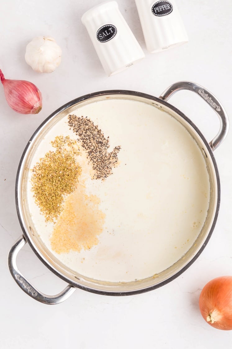 A pot of creamy white sauce with piles of black pepper, dried oregano, and garlic powder on top. Surrounding the pot are a shallot, a garlic head, an onion, and salt and pepper shakers on a white surface.