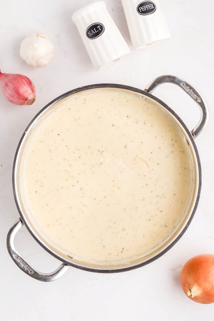 A pot filled with creamy white sauce sits on a white surface, surrounded by a red onion, a shallot, a garlic bulb, and salt and pepper shakers.