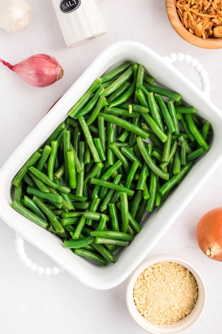 A white rectangular baking dish filled with fresh, cut green beans sits on a white surface, surrounded by a shallot, garlic, a salt shaker, a bowl of fried onions, and a bowl of breadcrumbs.