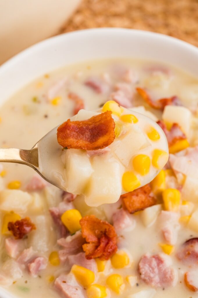 A close-up of a spoonful of creamy soup with diced potatoes, corn, ham, and a piece of crispy bacon, held above a bowl filled with the same soup.