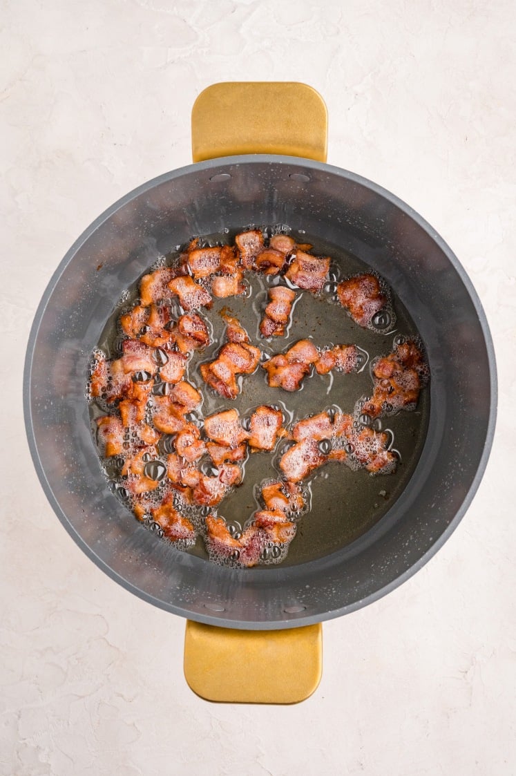 Crispy bacon pieces sizzling in hot oil inside a round black pot with gold handles, viewed from above on a light-colored surface.