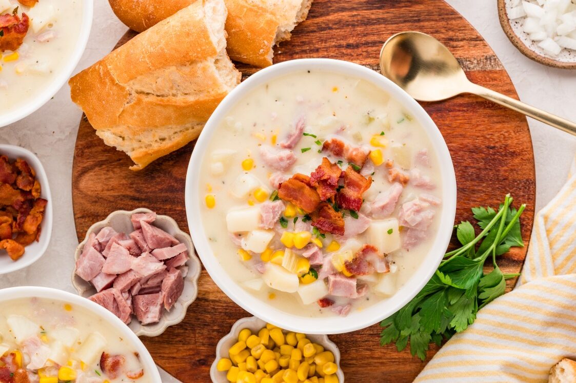 A bowl of creamy soup with ham, potatoes, corn, and bacon bits sits on a wooden board, surrounded by French bread, parsley, chopped ham, and corn, with a gold spoon nearby.