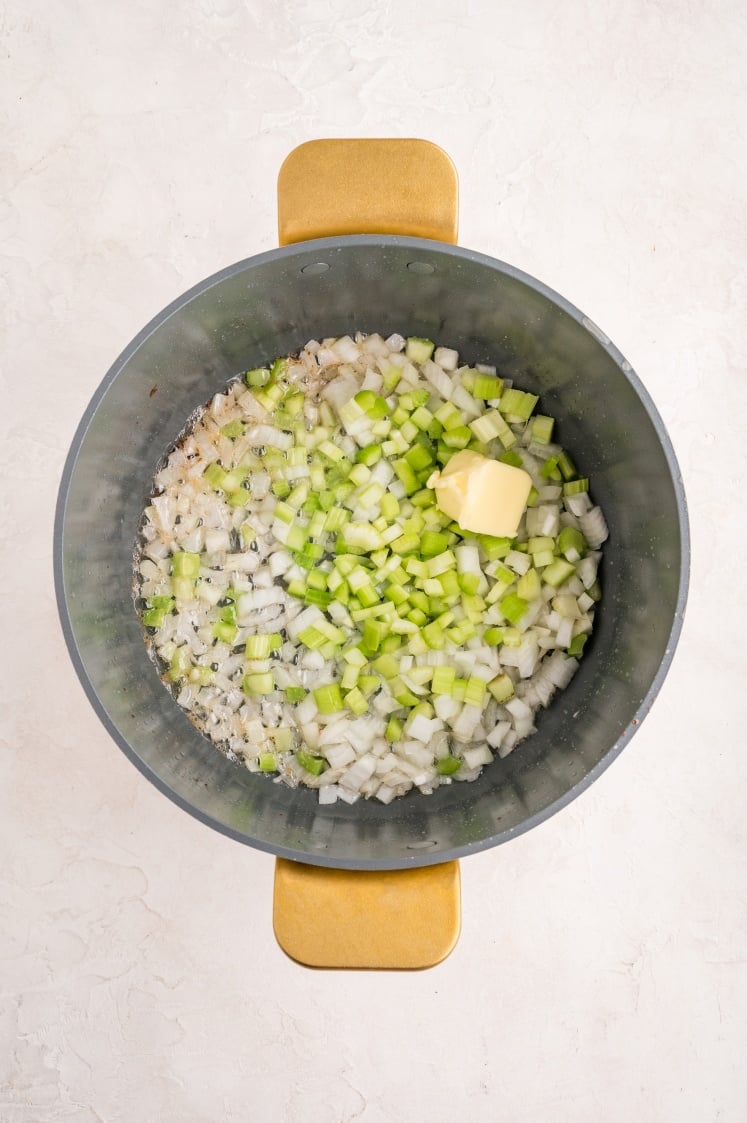 A pot containing chopped onions and celery with a pat of butter on top, ready to be cooked, viewed from above on a light-colored surface.