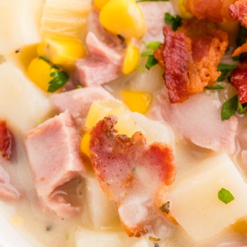 A close-up of a creamy soup featuring chunks of ham, potatoes, sweet corn, and crispy bacon, garnished with chopped parsley.