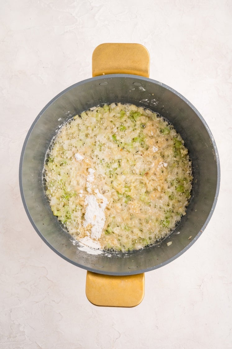 A top-down view of a pot with chopped onions and celery being sautéed in butter, with a small pile of flour sprinkled on top, all on a light-colored surface.