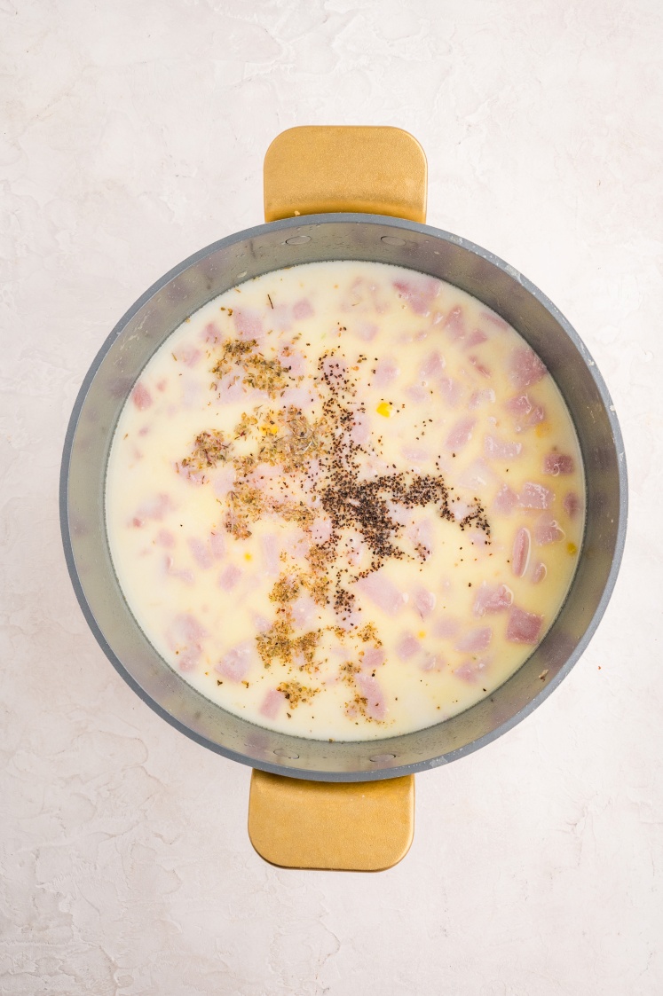 A pot filled with creamy soup containing chunks of ham, topped with black pepper and seasonings, sits on a light-colored surface.