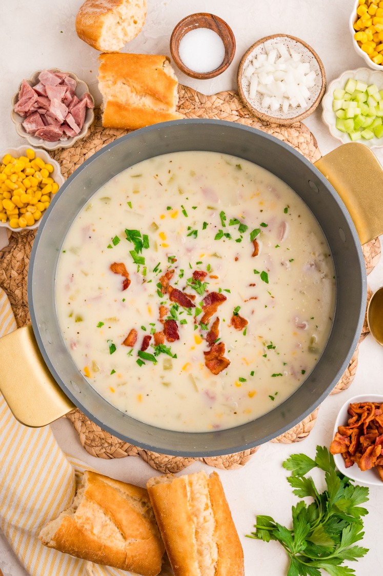 A pot of creamy soup garnished with parsley and bacon sits on a table, surrounded by ingredients like corn, ham, chopped onions, celery, extra bacon, and pieces of baguette bread.