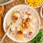 A bowl of creamy potato chowder with corn, bacon pieces, and potatoes, served with a spoon. A small bowl of corn, parsley, bread, and a pot of chowder are nearby on a woven mat.