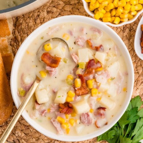 A bowl of creamy potato chowder with corn, bacon pieces, and potatoes, served with a spoon. A small bowl of corn, parsley, bread, and a pot of chowder are nearby on a woven mat.