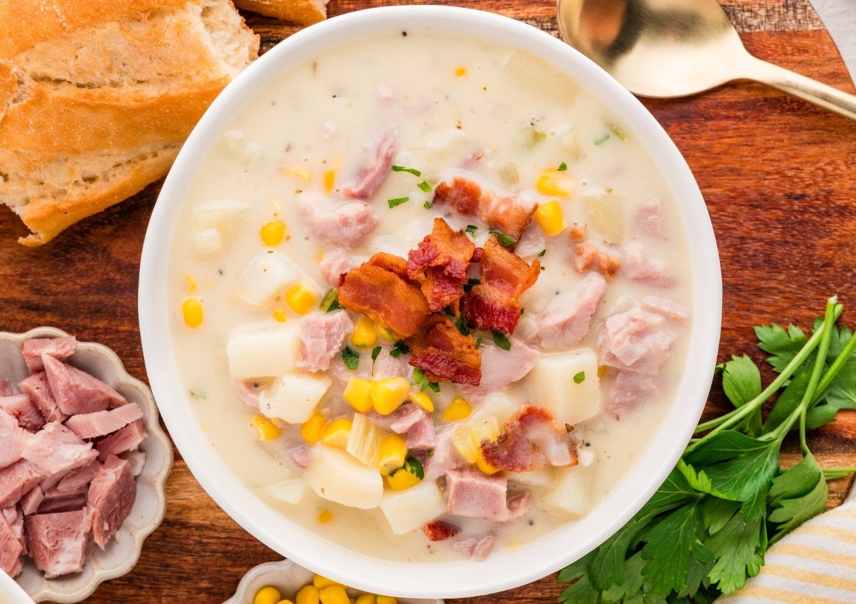 A bowl of creamy soup with diced ham, potatoes, corn, and topped with crispy bacon. Slices of bread, parsley, and extra ham are on the wooden surface nearby, along with a gold spoon.
