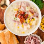 A bowl of ham and corn chowder topped with bacon bits, surrounded by bread, ham, corn, parsley, and a gold spoon on a wooden table. The text "Ham and Corn Chowder" is at the top.