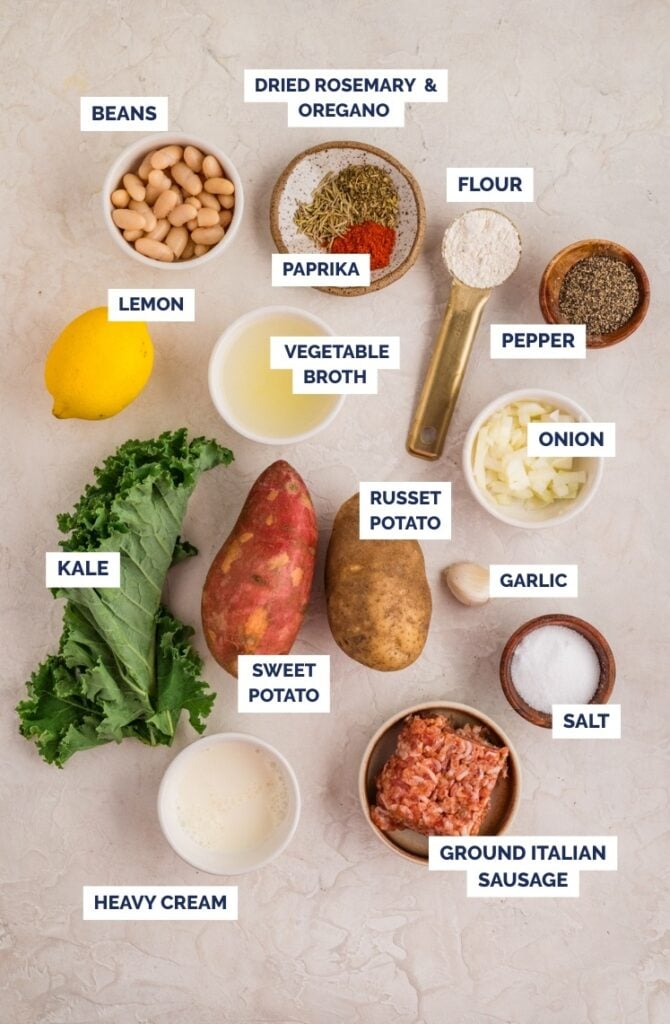 Ingredients for Kale and White Bean Sausage Soup arranged on a light surface, each labeled: beans, dried rosemary & oregano, flour, paprika, pepper, lemon, vegetable broth, onion, russet potato, sweet potato, garlic, salt, kale, sausage & heavy cream.