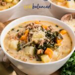 A hearty bowl of Kale and White Bean Sausage Soup with carrots, potatoes, and greens, topped with grated cheese, sits on a wooden board beside a spoon and fresh kale.