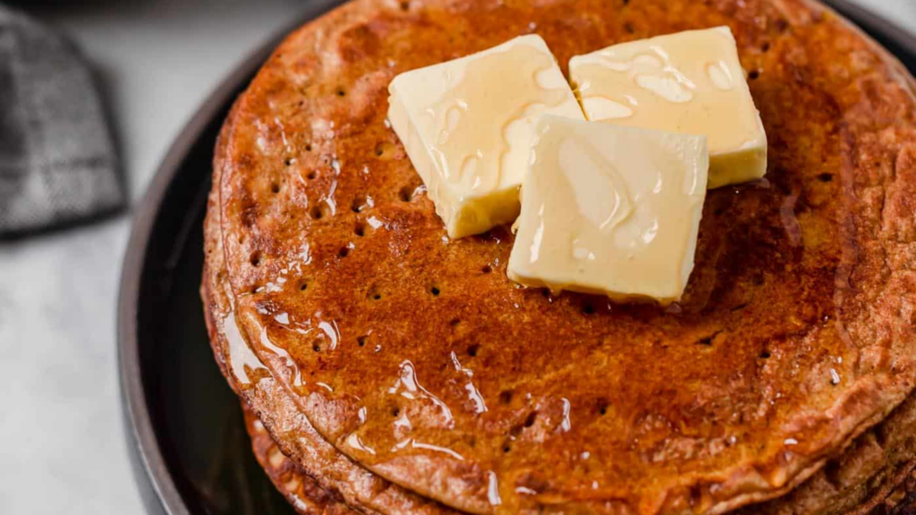 A stack of pumpkin pancakes topped with butter and syrup on a dark plate.