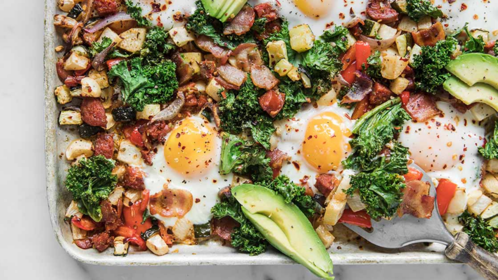 Overhead view of a baked breakfast hash on a sheet pan, featuring sunny-side-up eggs, chopped kale, avocado slices, red onion, and diced vegetables with a sprinkle of red pepper flakes.