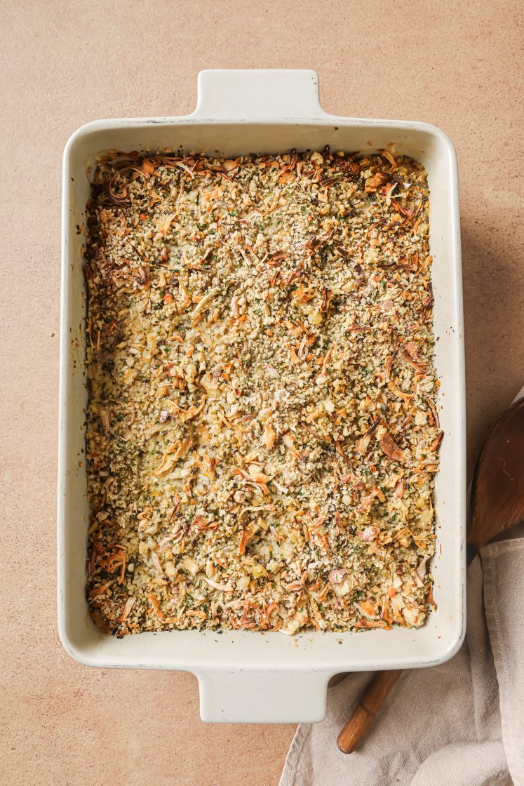 A rectangular white baking dish filled with a golden-brown, crumb-topped casserole sits on a light brown surface next to a beige cloth and a wooden serving spoon.