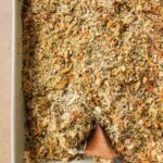 A baking dish filled with a golden, crumbly casserole topped with herbs and shredded vegetables. A wooden spoon rests inside the dish, revealing the creamy filling underneath the crispy topping.