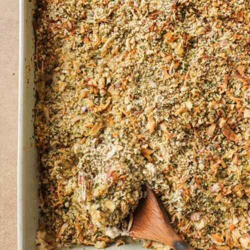 A baking dish filled with a golden, crumbly casserole topped with herbs and shredded vegetables. A wooden spoon rests inside the dish, revealing the creamy filling underneath the crispy topping.