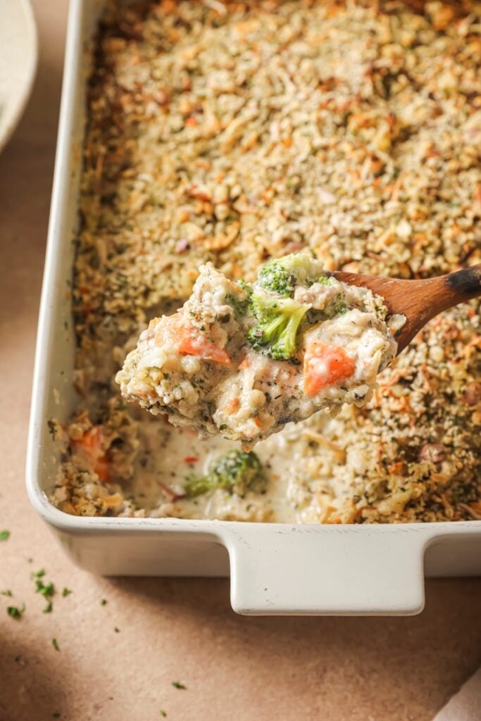 A close-up of a wooden spoon lifting a creamy vegetable casserole with broccoli and carrots from a white baking dish. The casserole has a crispy, golden topping.