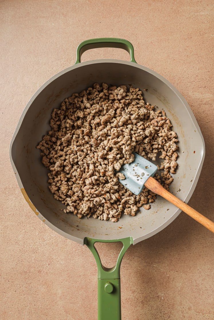 A green-handled skillet on a countertop contains cooked ground meat being stirred with a spatula that has a wooden handle and gray silicone head.