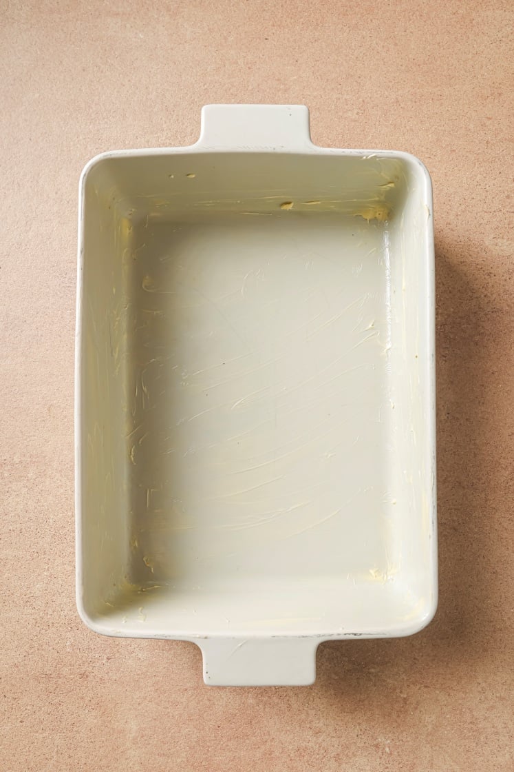 A rectangular white ceramic baking dish with handles sits on a tan surface. The inside of the dish is lightly coated with butter.