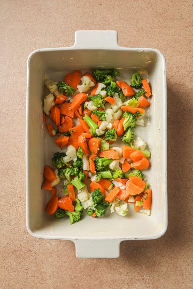 A white baking dish contains a mix of chopped broccoli, cauliflower, and carrots on a light brown countertop.
