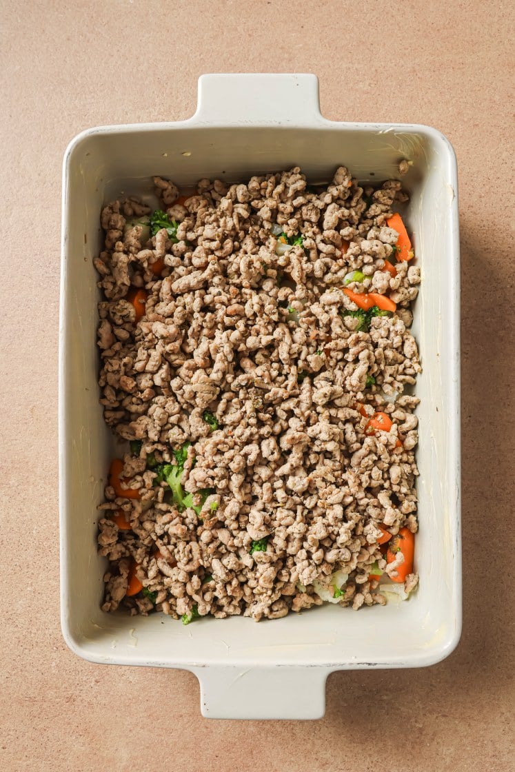 A rectangular baking dish filled with a layer of cooked ground meat mixed with vegetables such as broccoli and carrots, sitting on a light brown countertop.
