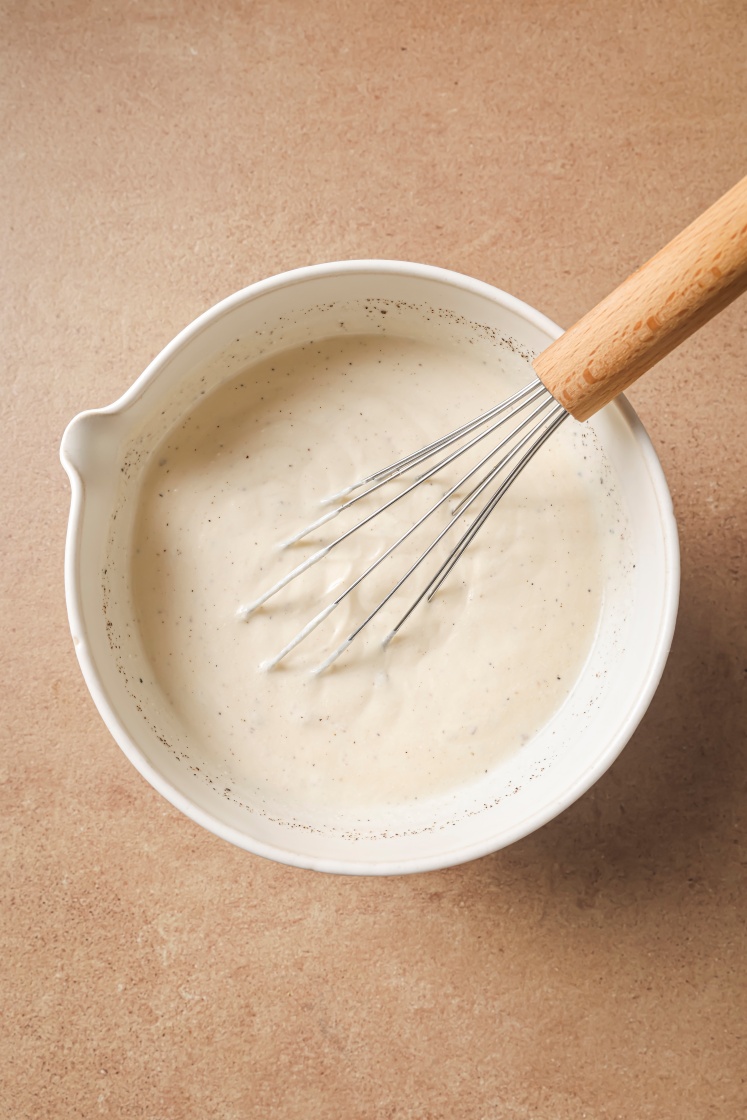 A white mixing bowl filled with creamy white sauce, with a metal whisk resting inside, on a light brown countertop.