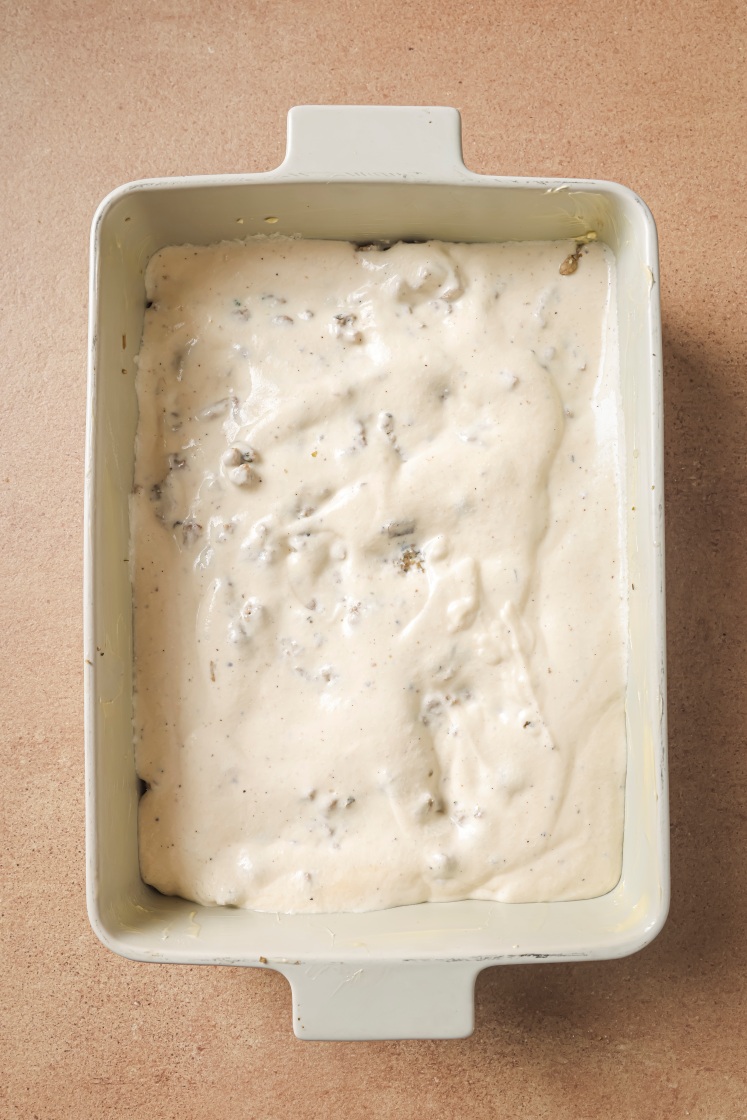 A rectangular baking dish filled with an uncooked, creamy white mixture with small chunks, likely a casserole or savory bake, sits on a tan countertop.