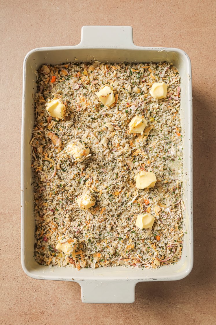 A rectangular baking dish filled with a breadcrumb and vegetable mixture, topped with several small dollops of butter, sits on a brown countertop.