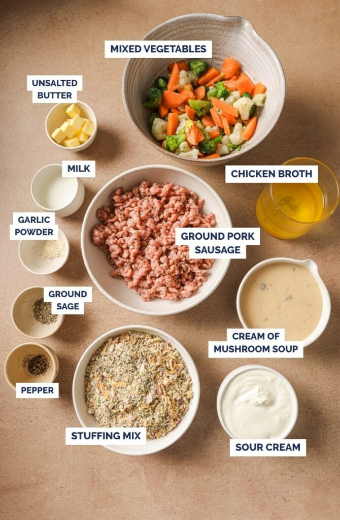 A top view of ingredients in separate bowls labeled: mixed vegetables, unsalted butter, milk, chicken broth, garlic powder, ground sage, pepper, ground pork sausage, cream of mushroom soup, stuffing mix, and sour cream.